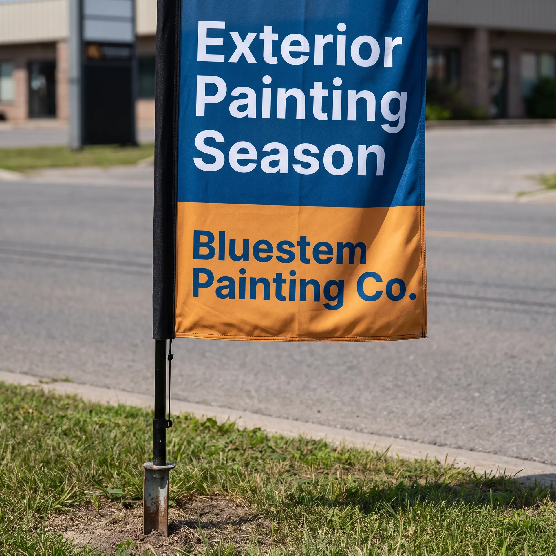 Printed feather flag on its flexible fibreglass pole with a ground stake, shown with a sample design: on a grassy verge beside a roadside business entrance