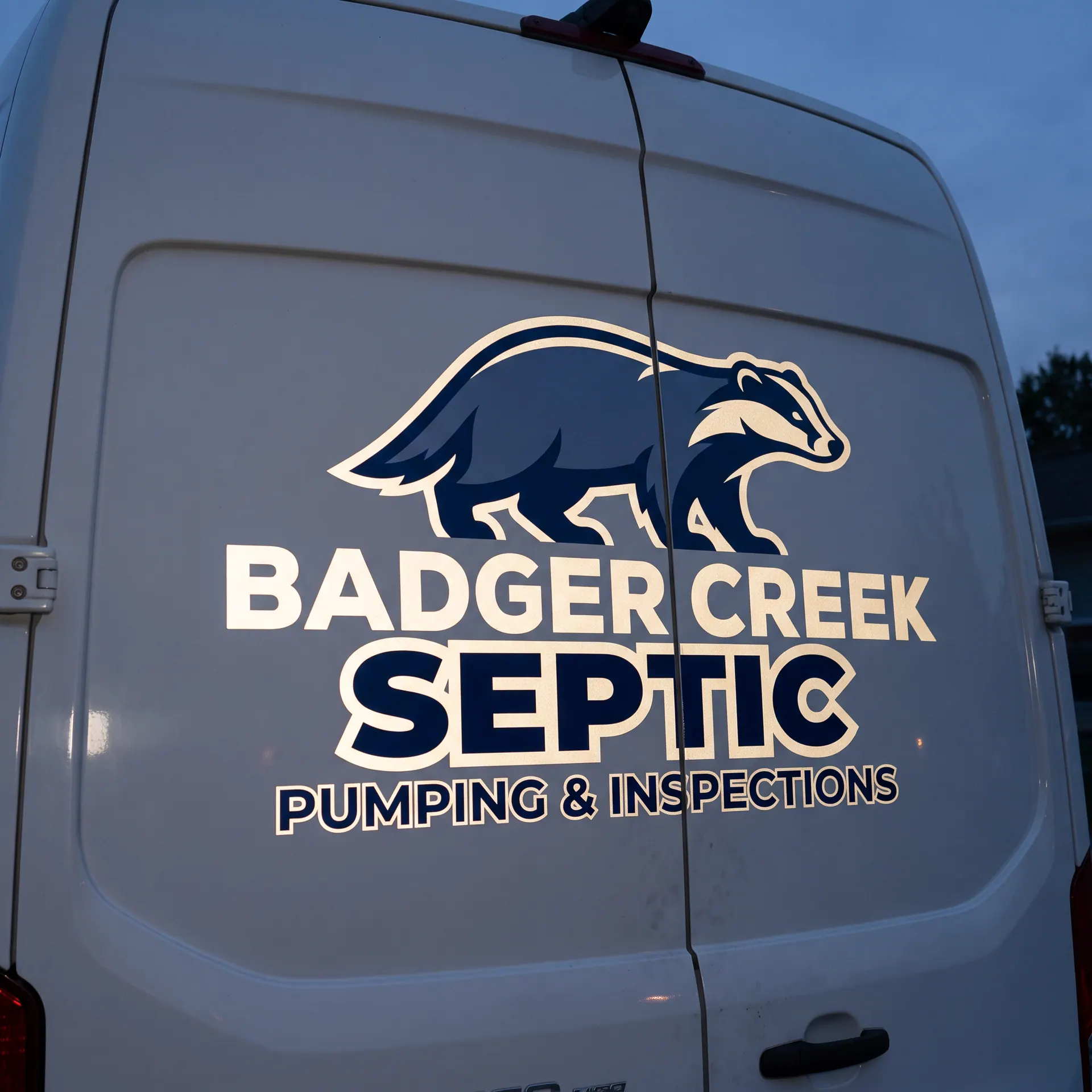 Printed reflective vinyl graphics applied directly to the painted metal of a van's rear doors, glowing brightly where headlights hit them; no panel, frame or grommets; the frame is cropped tight to the upper two thirds of the rear doors, so the bumper, licence plate and any manufacturer badge are out of frame, 24″ × 36″, shown with a sample design: on the rear doors of a white service van at dusk